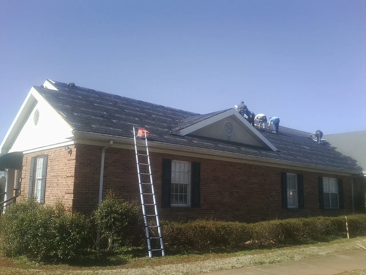 Crew tearing shingles off a brick ranch roof with a ladder set — Emergency Roof Repair work by our crew in Bohannon