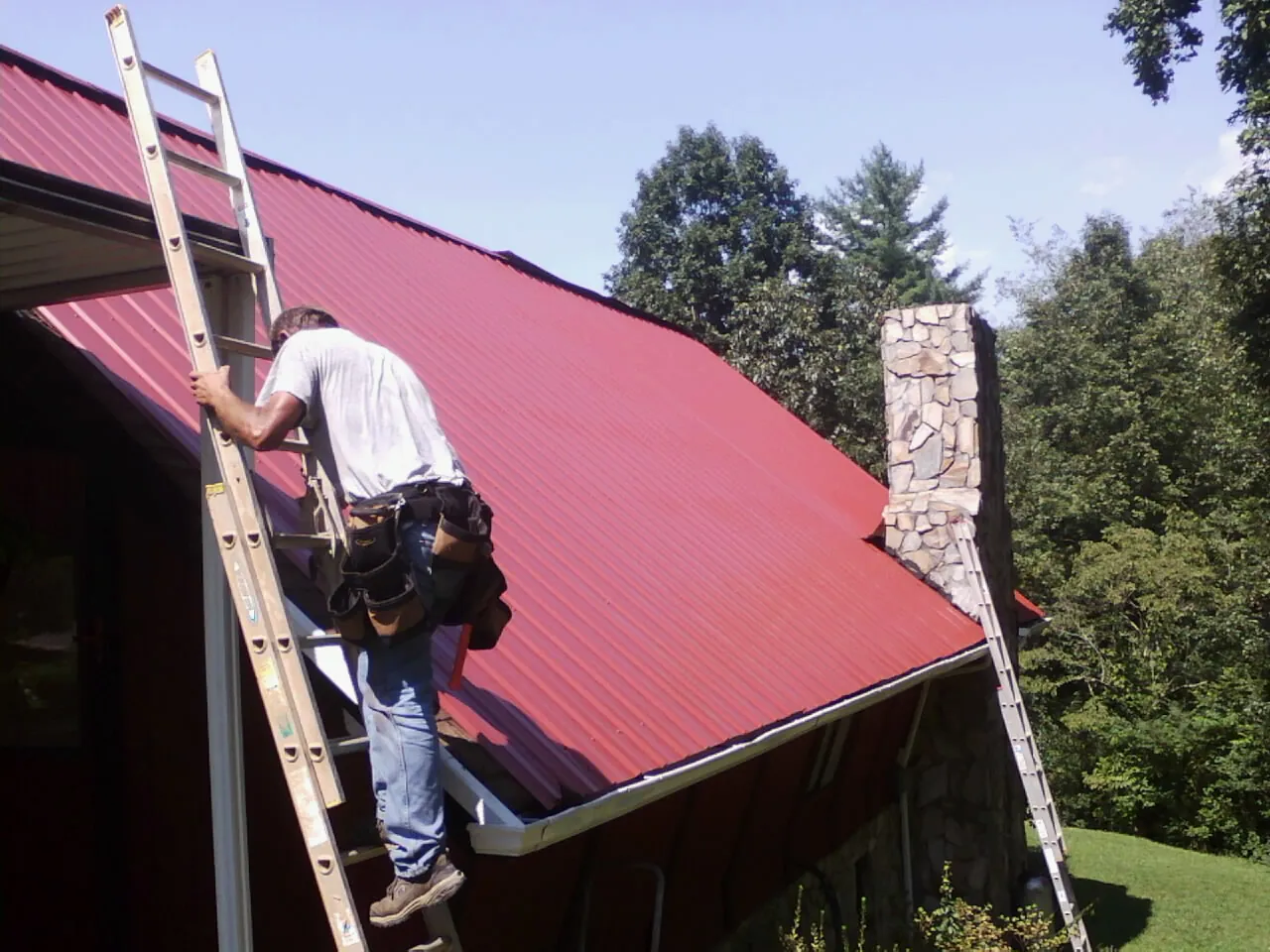 Roofer on a ladder beside a red metal roof and stone chimney — Soffit & Fascia Repair by Roofing Brothers in Bohannon